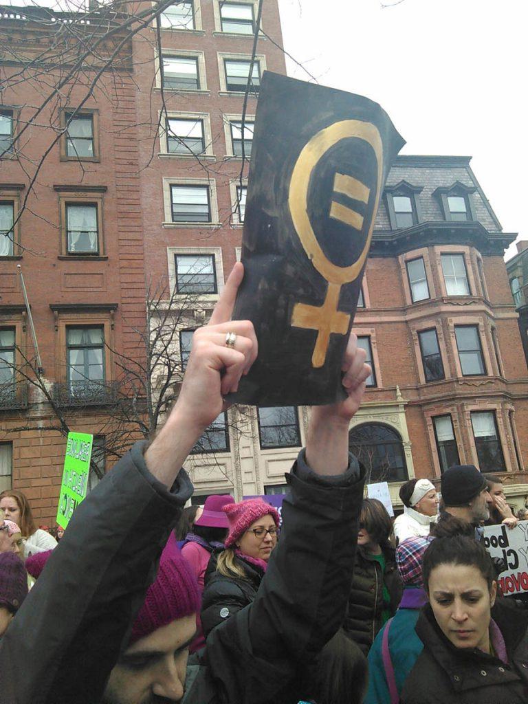 Photograph primarily depicts a sign held up by a protester in a crowd of people. The sign features a woman’s symbol with an equals sign.  Brick and sandstone buildings are in the background.  